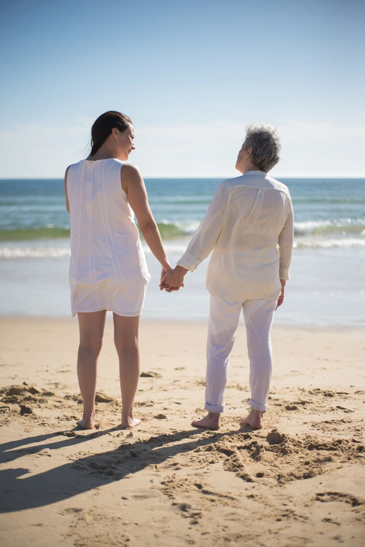 Adult daughter and mother holding hands on the beach while looking out at the shore.