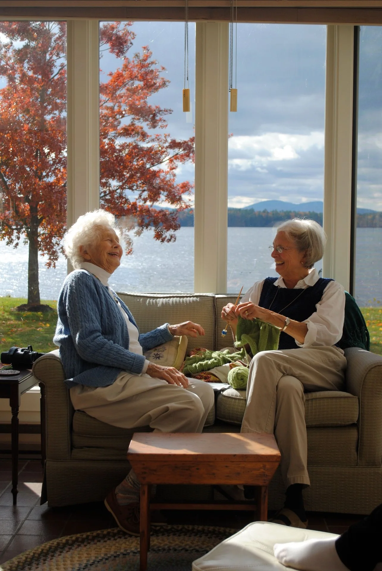 Two older women laughing on a couch in a lakehouse.