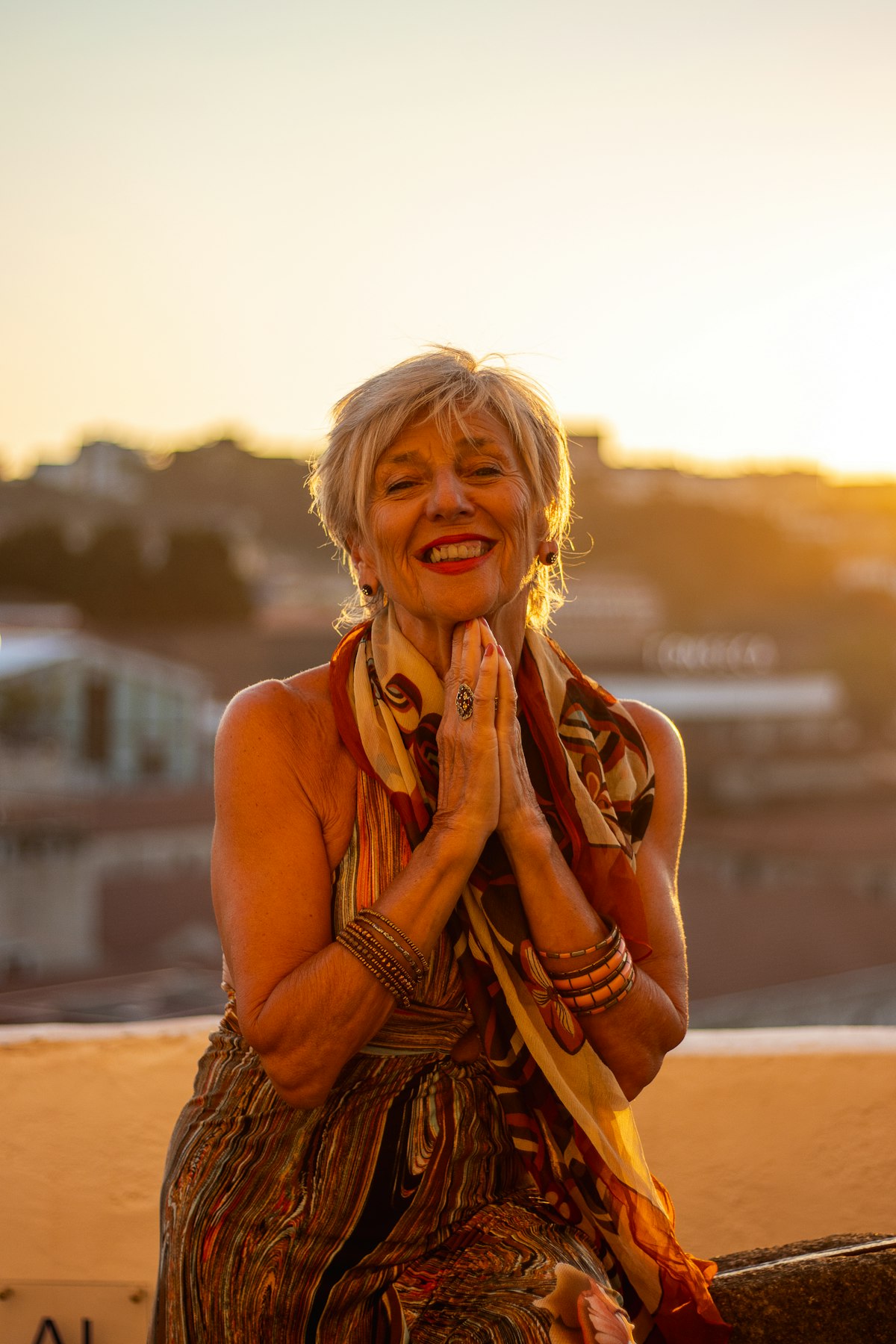 Woman with clasped hands smiling in front of a city skyline at sunset.