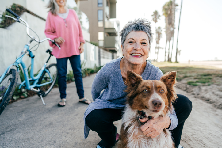 Older woman squatting and smiling while hugging her dog. Another older woman stands in the background next to a bike.