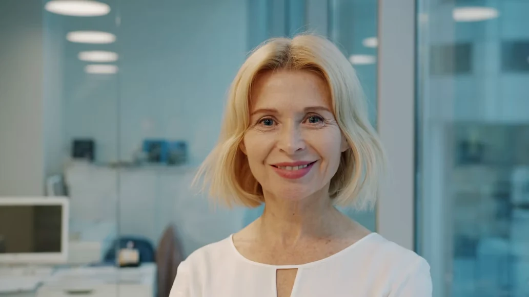 An older woman with blonde hair standing indoors, smiling